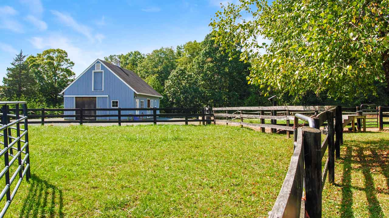 A well-maintained light blue barn with white trim sits within a fenced paddock under a bright blue sky. The barn features large double doors, a loft window, and multiple side windows. The surrounding area is lush with green grass, mature trees, and a sturdy black wooden fence enclosing the pasture. Sunlight filters through the leaves, casting dappled shadows on the ground.