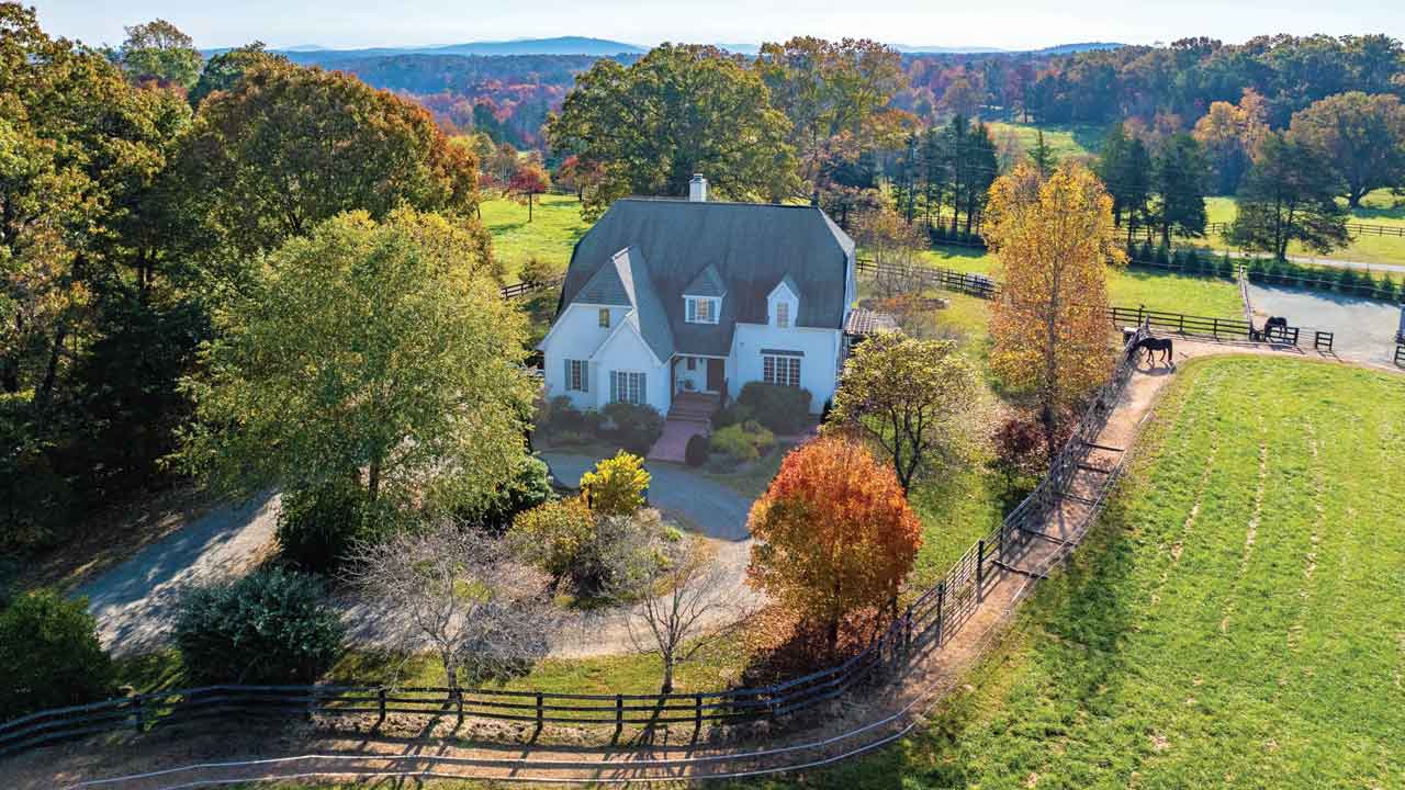 Aerial view of a charming white country home with a steep roof, surrounded by lush green fields, autumn-colored trees, and wooden fencing, with a horse grazing nearby. Rolling hills and forests stretch into the distance.