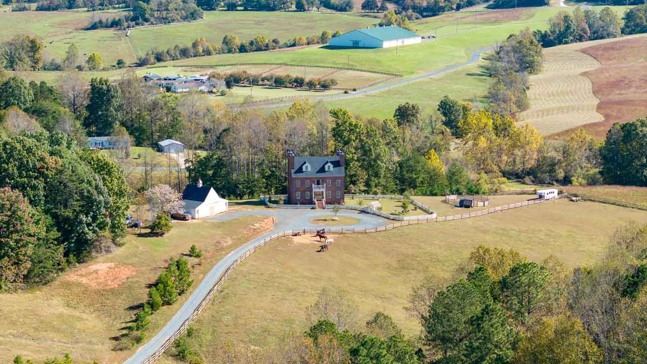 Aerial view of a grand brick estate with a long driveway, surrounded by rolling pastures and fenced horse paddocks. A horse is visible in the open field, with scenic farmland and a large barn in the background.