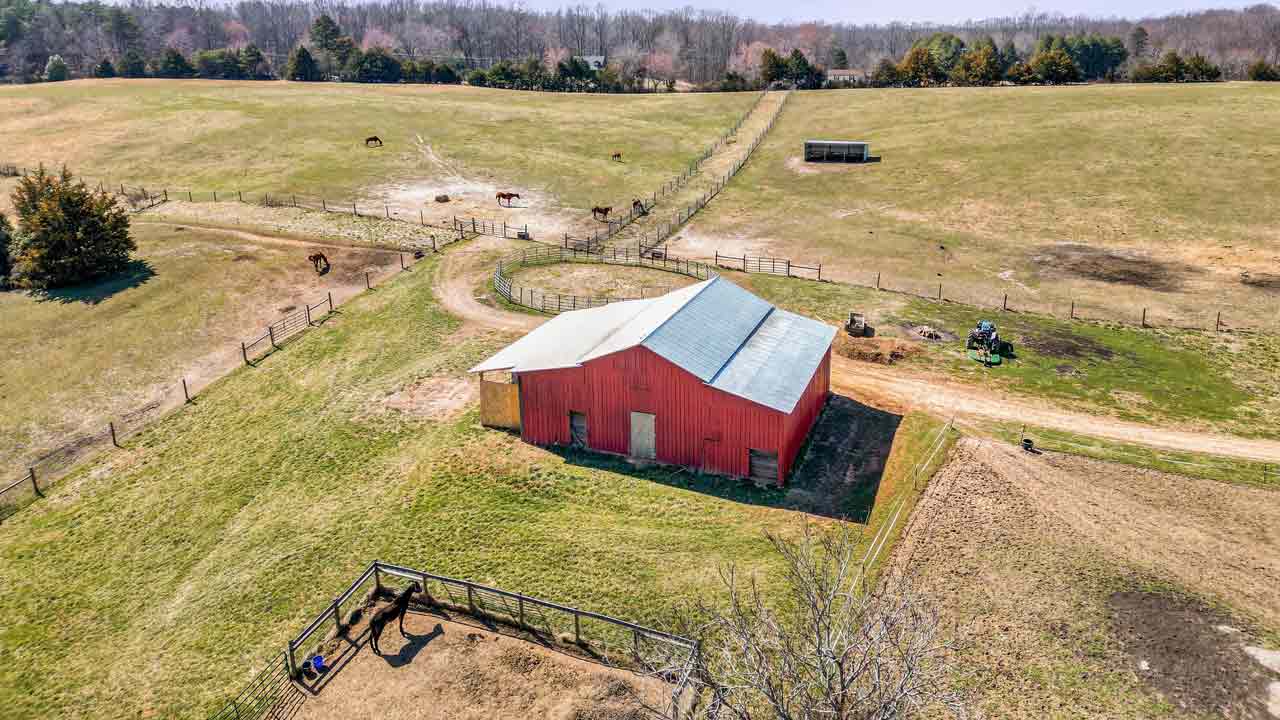 Aerial view of a charming white country home with a steep roof, surrounded by lush green fields, autumn-colored trees, and wooden fencing, with a horse grazing nearby. Rolling hills and forests stretch into the distance.