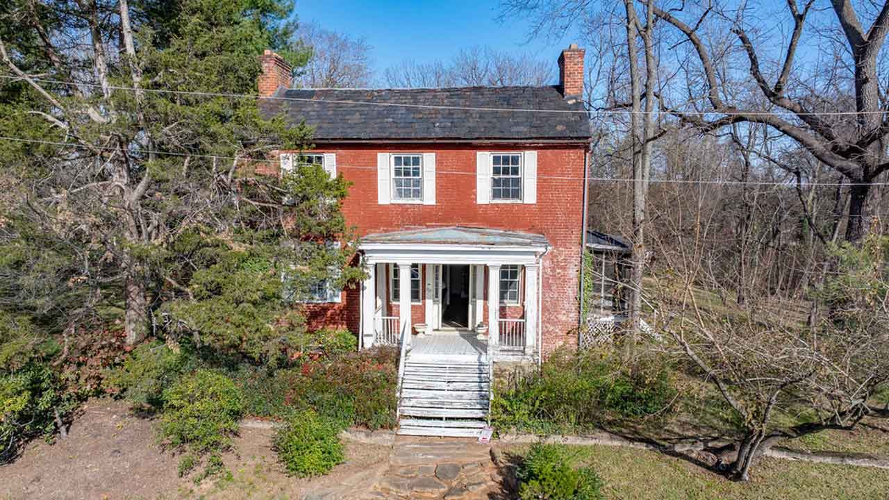 Front view of a historic red brick home with a white porch, framed by tall trees. The house features a classic design with large windows and a stone walkway leading to the entrance. The surrounding landscape is wooded and bare, reflecting a late autumn or early winter setting.