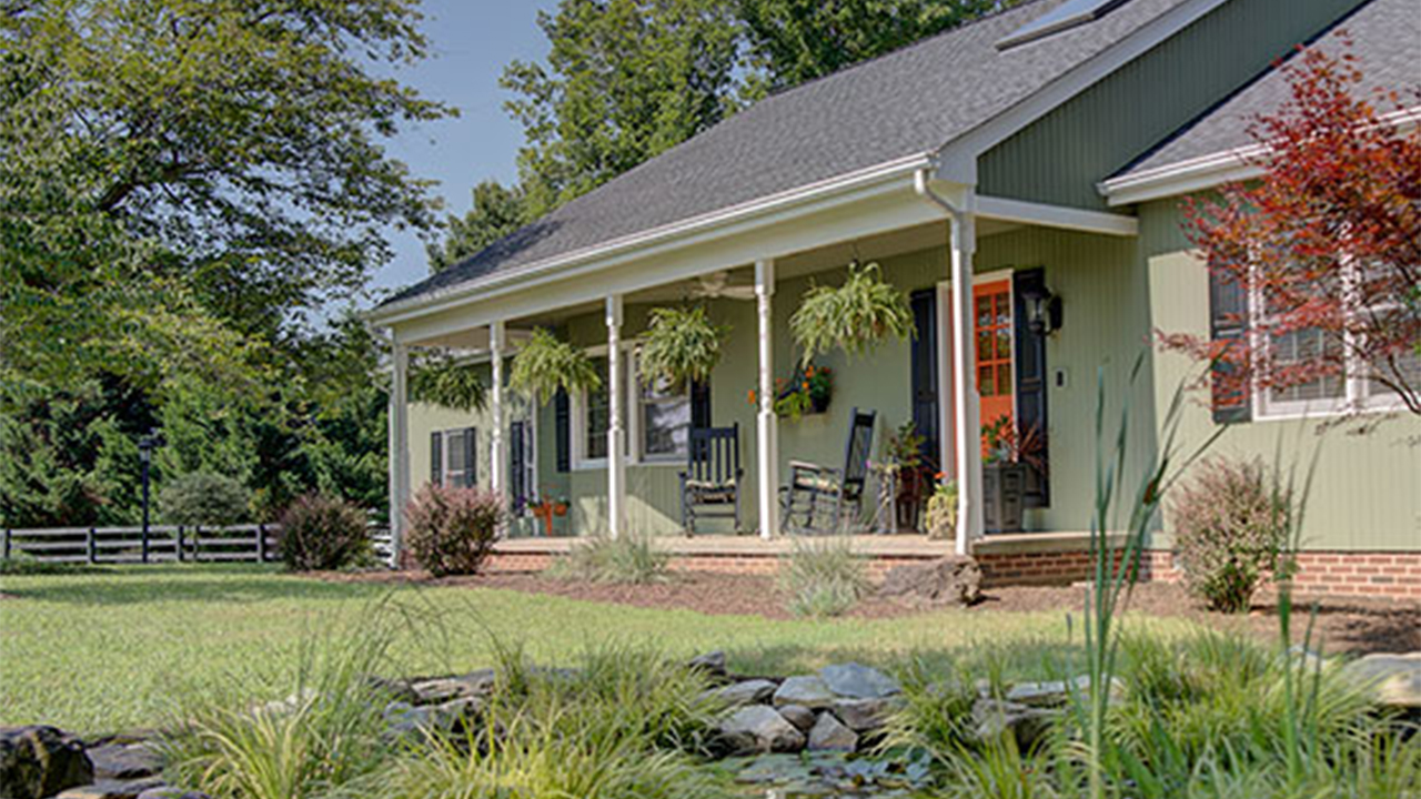 A charming country home with soft green siding, black shutters, and a welcoming front porch adorned with hanging ferns and rocking chairs. The house features a vibrant orange front door, white columns supporting the porch roof, and brick skirting along the foundation. The well-maintained landscaping includes ornamental grasses, flowering plants, and a small rock-lined pond in the foreground, creating a peaceful rural retreat.