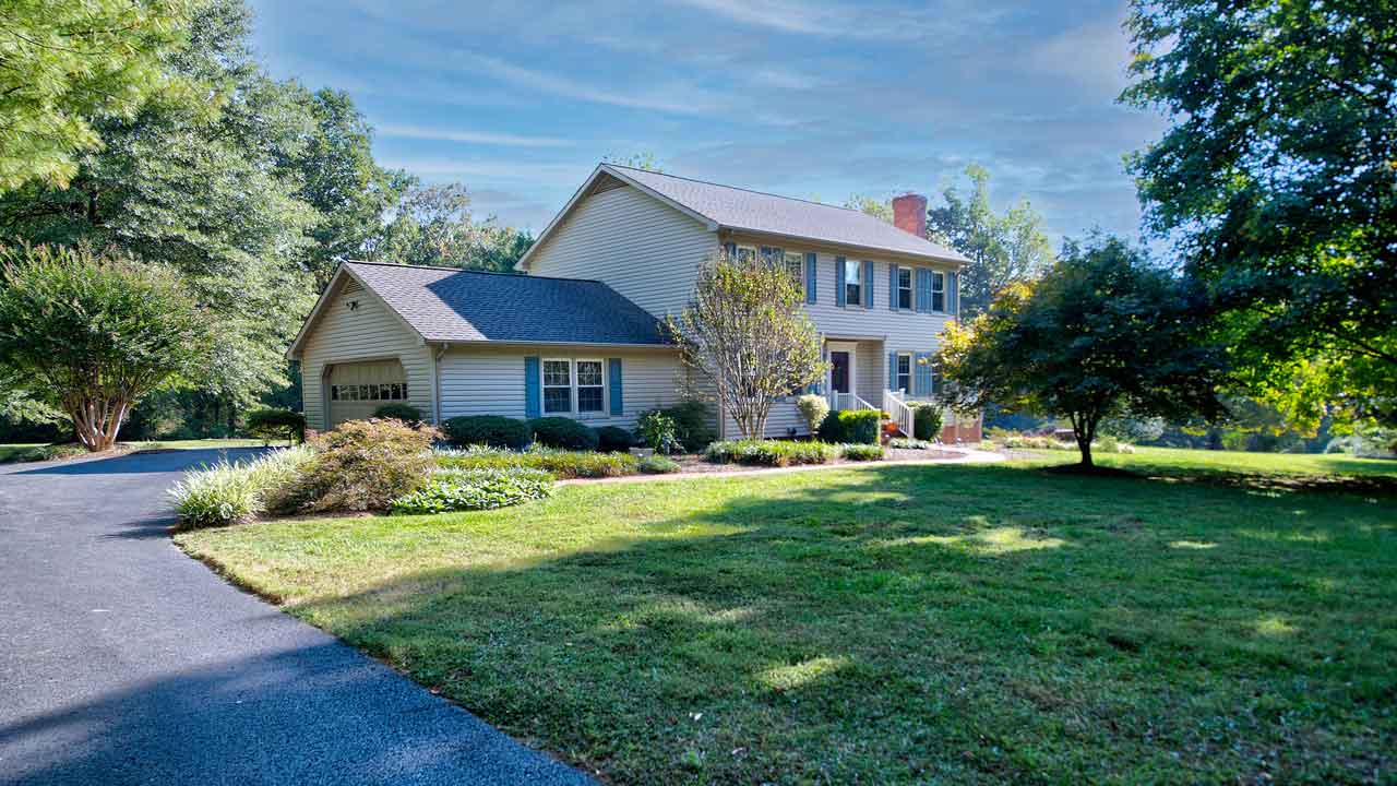 A beautifully maintained two-story suburban home with beige siding, black shutters, and a brick chimney, nestled in a lush green landscape. The property features a welcoming front porch, an attached garage with a gabled roof, and a curved asphalt driveway leading to the entrance. The manicured lawn is accented with mature trees, flowering shrubs, and neatly landscaped garden beds, creating a peaceful and inviting atmosphere.