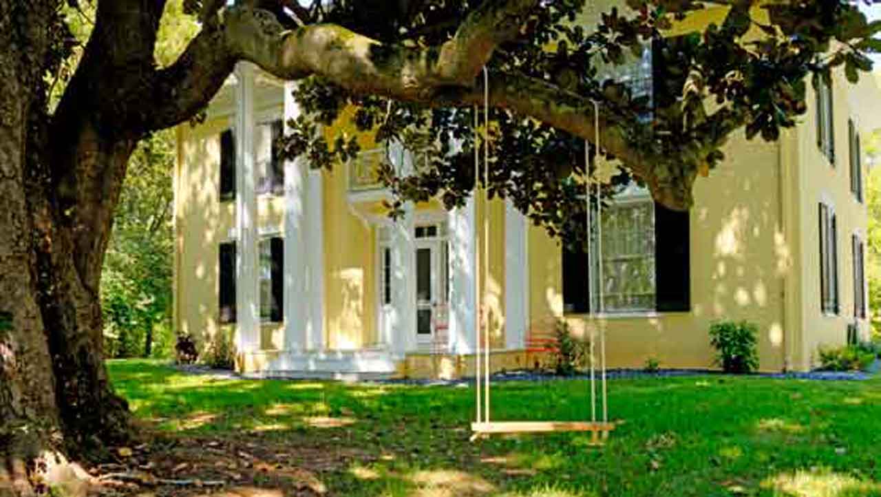 Charming yellow historic home with white columns and black shutters, nestled among lush greenery. A wooden tree swing hangs from a large, sprawling tree in the foreground, creating a picturesque and inviting scene.