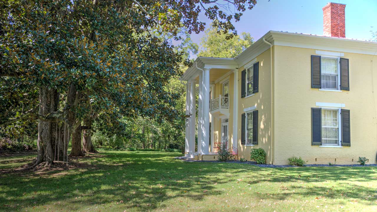 A distinguished two-story Southern-style residence with a soft yellow brick exterior, accented by black shutters and a striking columned portico. The entrance boasts a second-story balcony with an ornate railing, while a prominent red brick chimney adds to its classic charm. Nestled amid a lush green lawn and framed by mature trees, the home exudes timeless elegance and tranquility.