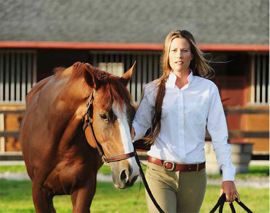 Equestrian woman leading a chestnut horse with a white blaze, dressed in a white shirt and beige riding pants, with a stable and fenced pasture in the background.