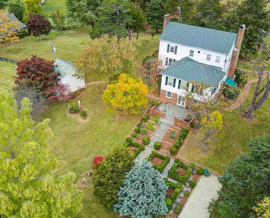 Aerial view of a charming two-story white farmhouse with a green metal roof, surrounded by colorful trees, manicured gardens, and a spacious lawn, with a nearby silo and scenic countryside setting.