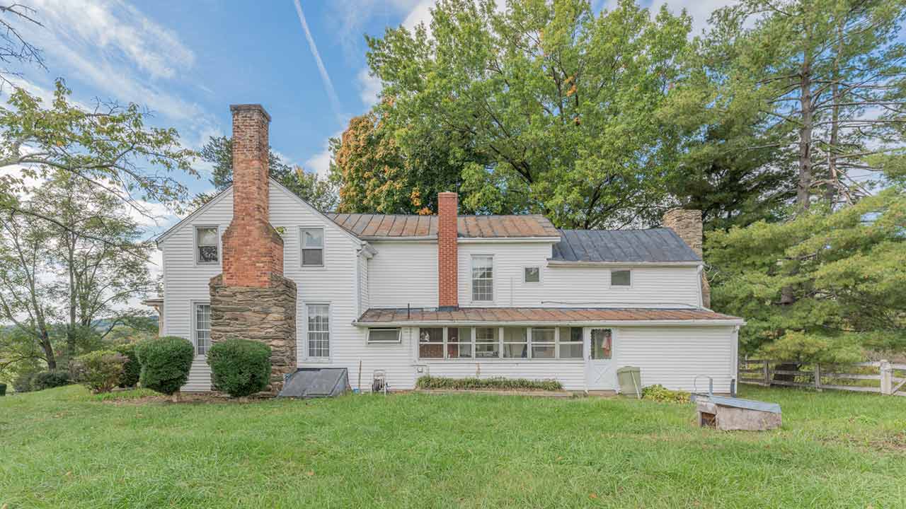 Charming historic farmhouse with white siding, a rustic metal roof, and a prominent stone chimney, nestled among mature trees. The home exudes country charm with its timeless architecture and tranquil rural setting.