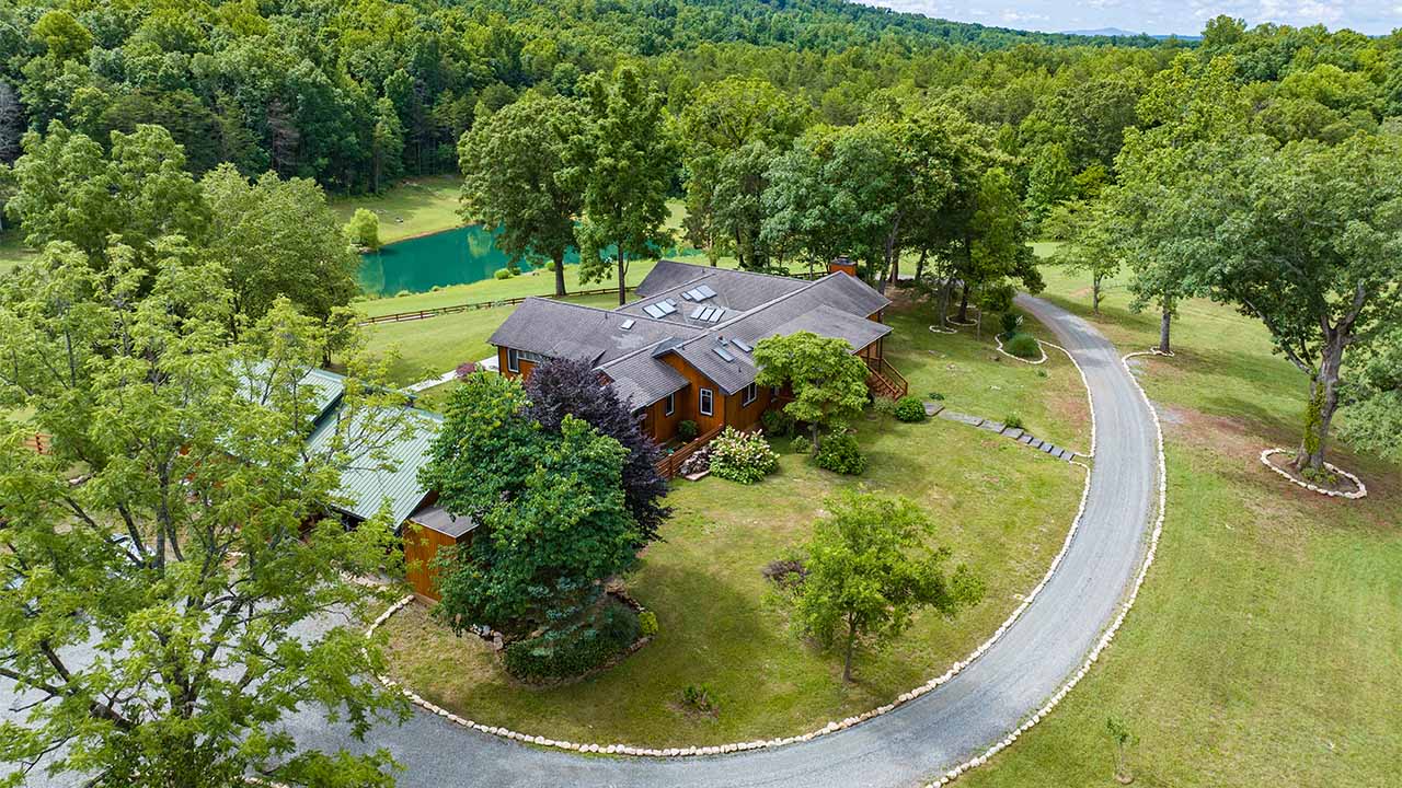 Aerial view of a custom-built rustic home with a dark roof, multiple skylights, and warm wood siding, surrounded by lush greenery. A curved gravel driveway leads to the house, which is nestled among mature trees and well-manicured landscaping. A picturesque pond with a wooden fence borders the property, set against a backdrop of rolling hills and dense forest, offering a serene and private retreat.