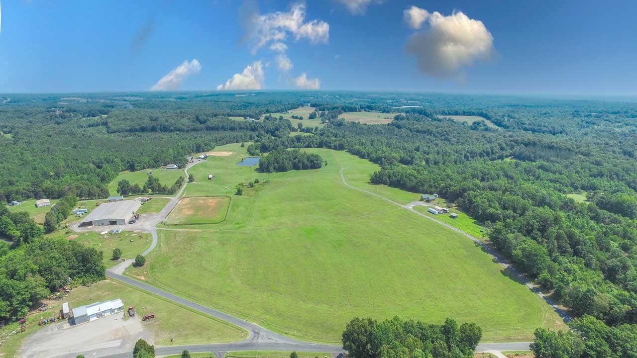 Aerial view of a sprawling equestrian facility with large open pastures, a training arena, multiple barns, and a scenic pond. The expansive property is surrounded by lush greenery and rolling countryside under a bright blue sky.