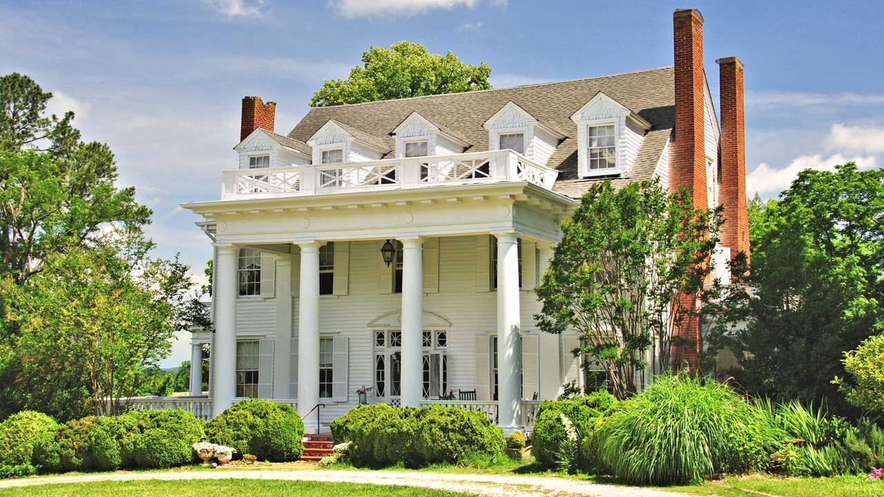 Grand Southern plantation-style home with a white columned portico, dormer windows, and multiple brick chimneys. The stately mansion features a second-story balcony with decorative railings, surrounded by lush greenery and manicured landscaping under a bright blue sky.