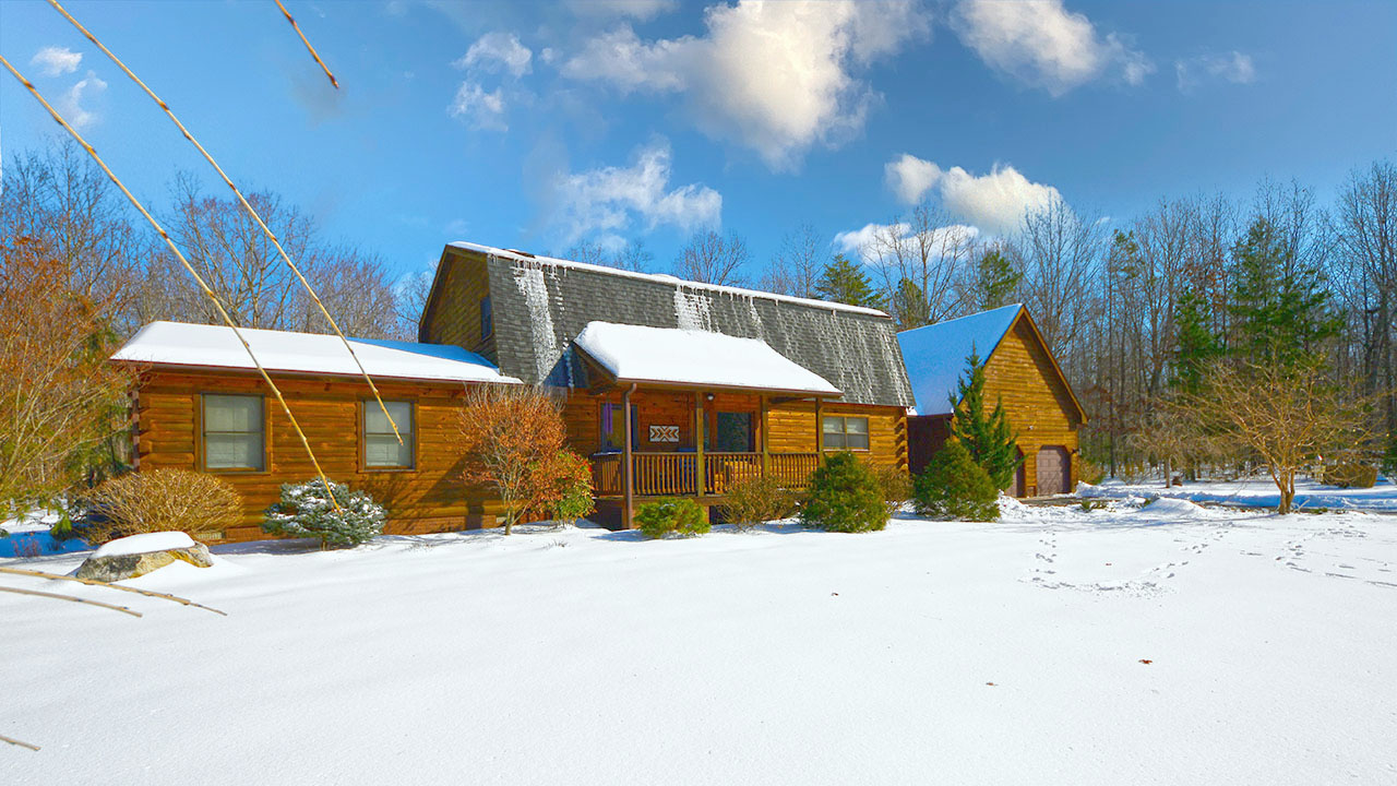 Charming log cabin home with a gambrel roof, covered porch, and detached garage, set in a serene winter landscape with fresh snow. Icicles hang from the roof as the rustic retreat sits under a bright blue sky with scattered clouds.