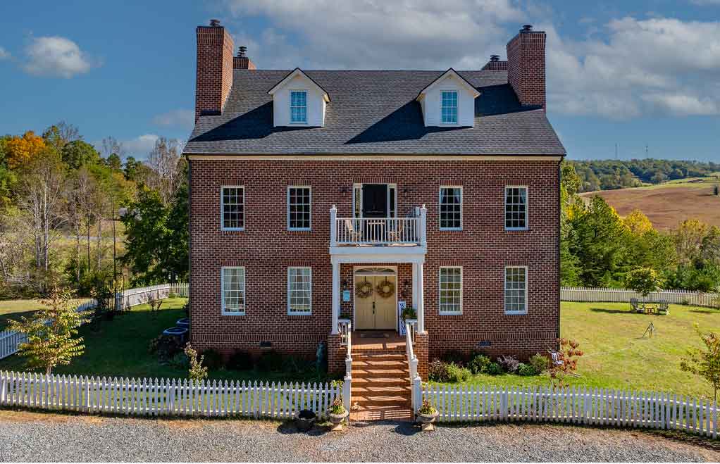 Elegant red brick Federal-style home with symmetrical windows, twin chimneys, dormer windows, and a central balcony above a columned entryway, surrounded by a white picket fence and set against a backdrop of greenery and clear blue skies.