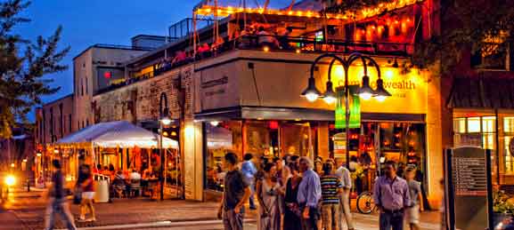 A lively evening scene at a downtown area with people walking and socializing outside a warmly lit restaurant. The building features a rooftop bar with string lights and an outdoor dining area with a canopy. The street is illuminated by vintage-style street lamps, creating a vibrant and inviting atmosphere.