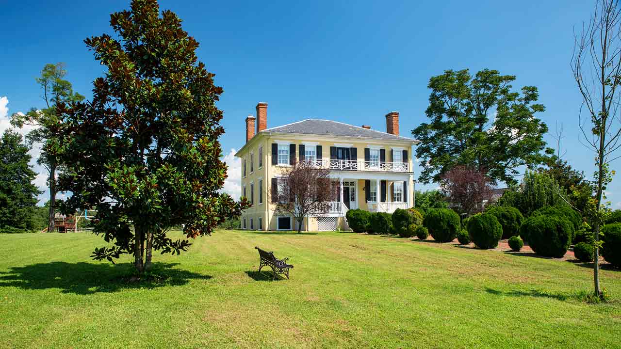 Stately Southern plantation-style home with a pale yellow brick exterior, black shutters, and a grand two-story balcony, surrounded by lush greenery and mature trees under a bright blue sky.