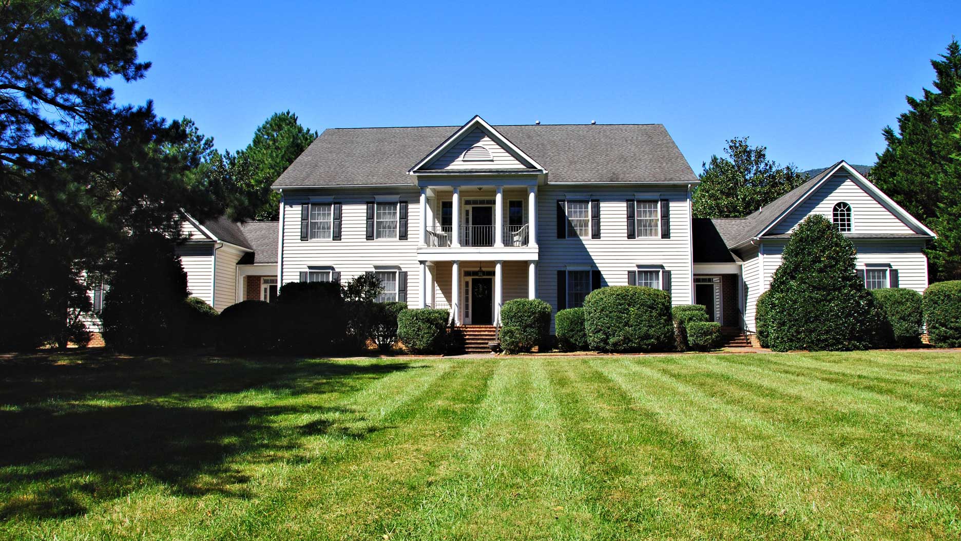 Elegant Colonial-style home in Virginia with a classic white exterior, black shutters, a grand columned entrance, and a second-story balcony, set on a beautifully manicured lawn with mature landscaping.