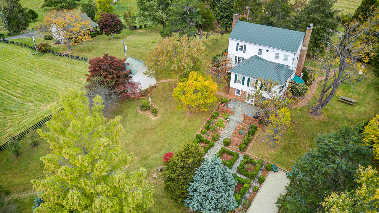 Aerial view of a historic white farmhouse with a green metal roof, surrounded by beautifully landscaped gardens, mature trees, and open pasture. A terraced stone pathway leads to the entrance, highlighting the estate's charm and country elegance.