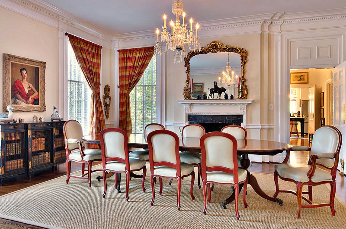 Elegant dining room with a polished wood table surrounded by vintage-style chairs featuring red frames and cream upholstery, a grand chandelier, ornate mirror above a white mantel, and rich plaid curtains framing tall windows, creating a classic and sophisticated ambiance.