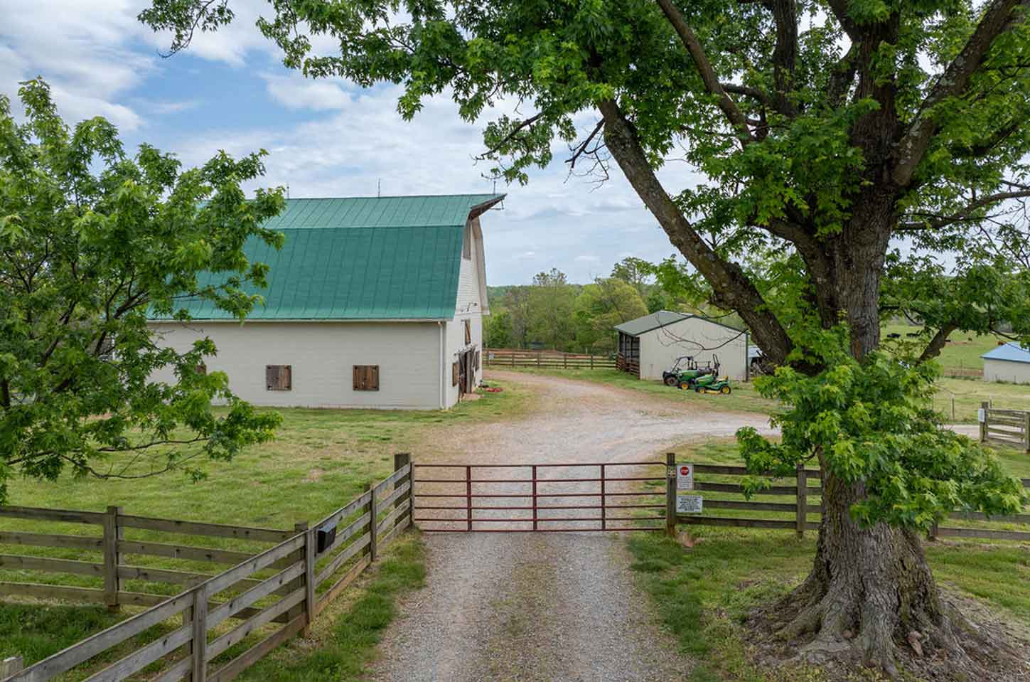 Scenic horse farm view featuring a white barn with a green roof, surrounded by a gravel driveway, wooden fences, and lush greenery, with a large tree in the foreground and additional farm structures visible in the background under a partly cloudy sky.