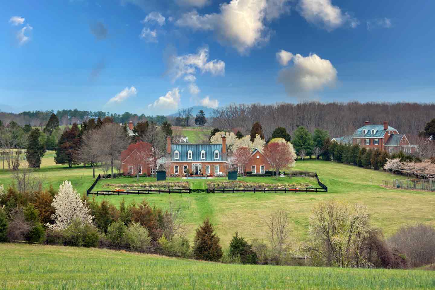 A stunning brick estate surrounded by lush greenery and blossoming trees, featuring a manicured lawn, a black wooden fence, and a backdrop of rolling hills under a vibrant blue sky with scattered clouds.