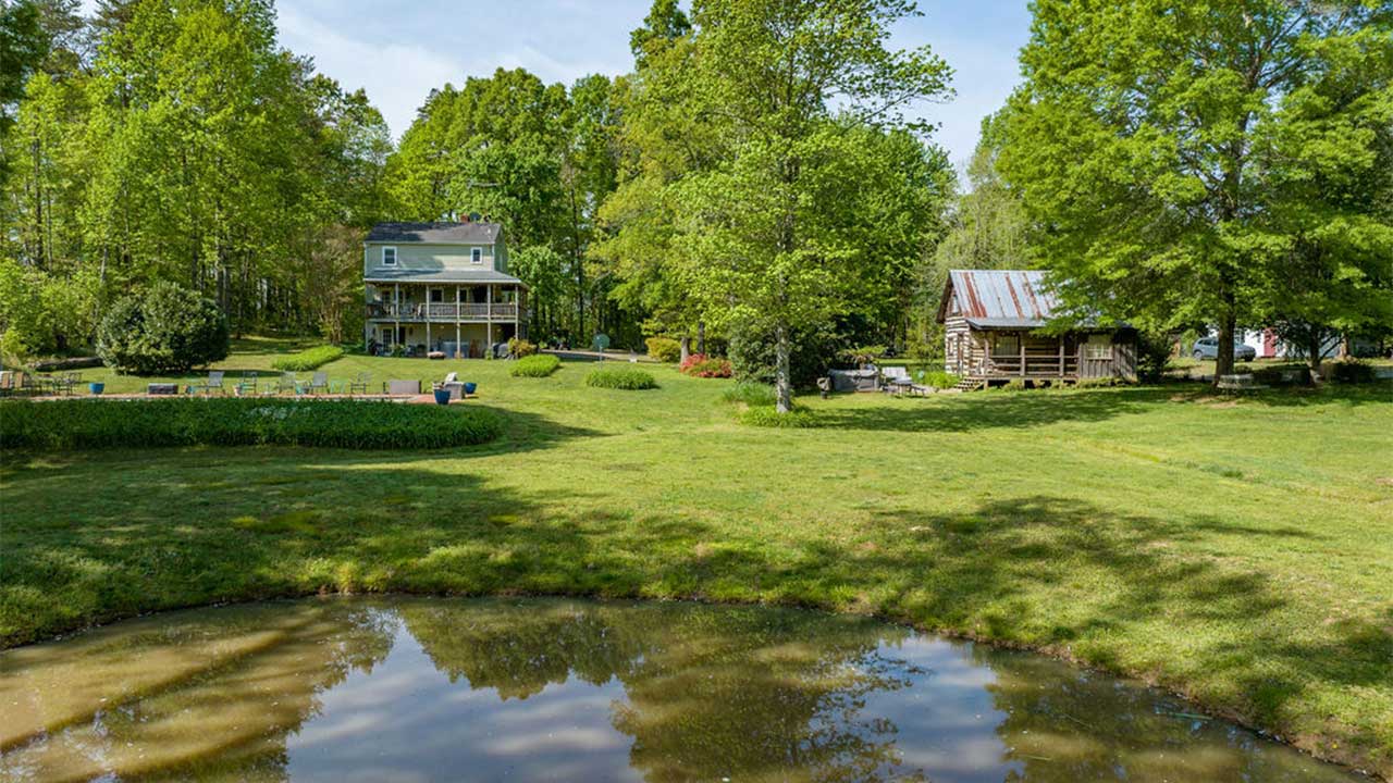 Scenic country property in Albemarle County, VA, featuring a charming two-story farmhouse with a wraparound porch, a rustic log cabin, and a tranquil pond, surrounded by lush greenery and mature trees.