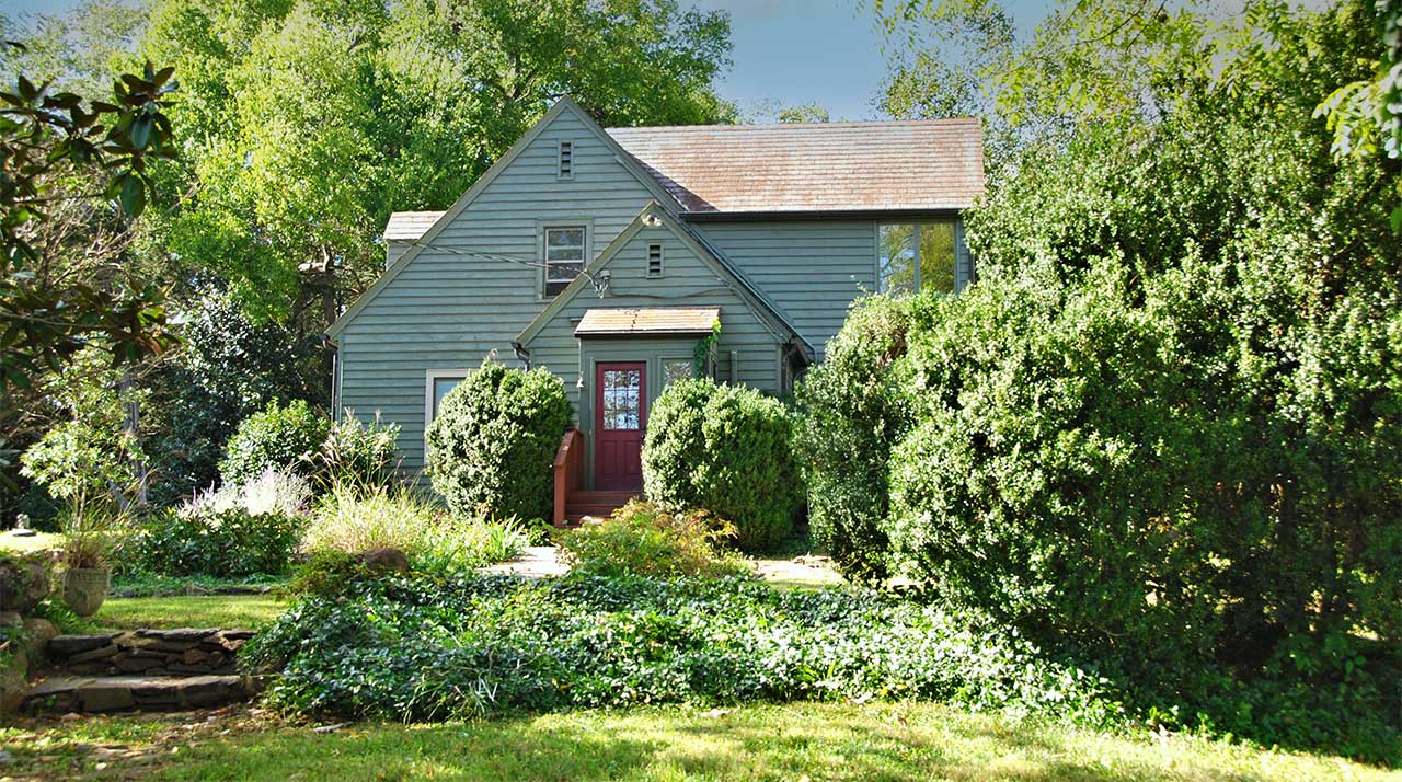 A rustic farmhouse with white siding, multiple chimneys, and a mix of metal and shingle roofing, set against a backdrop of mature trees and a blue sky. The home features a large stone chimney on one side, a long enclosed porch, and a basement entry with cellar doors. The surrounding lawn is lush and green, adding to the property's peaceful rural charm.